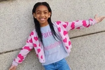 Paris smiling with her arms out, wearing a pink heart cardigan over a Paris Cares Foundation shirt